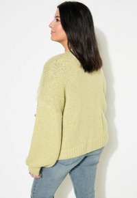 Suéter de punto verde claro con mangas oversized, dobladillo acanalado y textura suave. Lleva puesto sobre unos vaqueros, visto desde atrás.