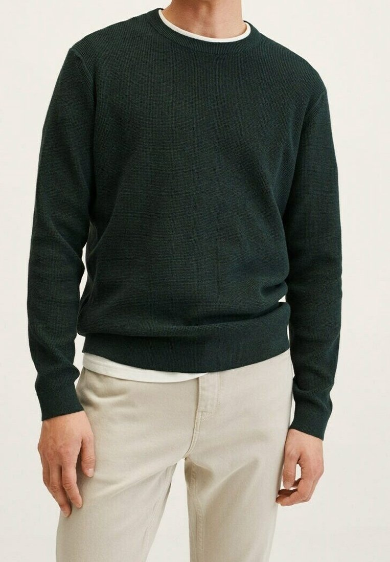Homme portant un pull vert foncé à col rond par-dessus un maillot blanc, associé à un pantalon beige clair, debout devant un fond uni.