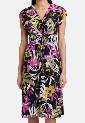 Kobieta ubrana w czarną sukienkę do kolan z marszczeniem z przodu i dużym kwiatowym wzorem w odcieniach fioletu, żółci, bieli i beżu.