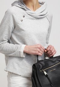 Persona che indossa un hoodie grigio chiaro con bottoni decorativi, mentre tiene una borsa in pelle nera con tasca frontale con zip e dettagli dorati.