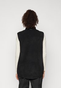 Schwarze Fleece-Weste mit hohem Kragen, ärmellosem Design, strukturiertem Oberflächen und abgerundetem Saum, kombiniert mit einem hellen Langarmoberteil darunter.