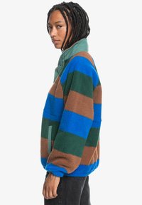 Veste en polaire avec des rayures vertes, bleues, marron et vert foncé. Elle présente un col haut et des poches latérales. Tissu doux et texturé.