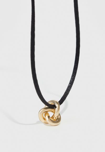 Pendentif en métal doré avec un design entrelacé sur un collier en corde noire fine. Surface lisse avec une finition polie, style minimaliste.