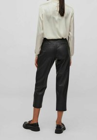 Pantalon en cuir noir à texture lisse, longueur raccourcie, avec des poches arrière. Associé à un chemisier en soie légère avec un col montant.