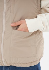 Béžová vypchávková vesta so zipsom, obsahujúca bočnú kapsu a rebrované manžety. Hladká textura s jemnými švovými detailmi.
