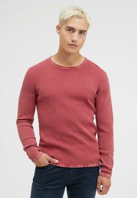 Rotes, strukturiertes Langarmhemd mit Rundhalsausschnitt und feinen Rippungen, kombiniert mit dunklen Jeans, auf einem neutralen Hintergrund präsentiert.