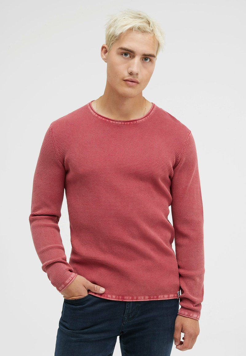 Rotes, strukturiertes Langarmhemd mit Rundhalsausschnitt und feinen Rippungen, kombiniert mit dunklen Jeans, auf einem neutralen Hintergrund präsentiert.