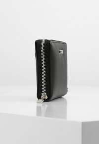 Cartera de cuero negra con un acabado texturizado, que cuenta con un cierre de cremallera plateado a lo largo de un lado y un pequeño acento de logo metálico.