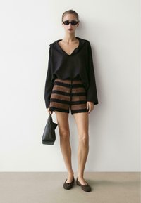 Vrouw met een zwarte oversized blouse, bruin-zwart gestreepte gehaakte shorts, zwarte platte schoenen, zonnebril, en een zwarte handtas, staand tegen een witte muur.