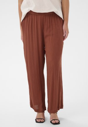 Femme portant un pantalon ample de couleur rouille texturé avec une taille élastique et des sandales noires à bouts ouverts, debout devant un fond uni.