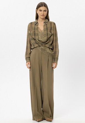 Woman with long brown hair wears loose olive green pants, olive lace camisole, and brown plaid button-up shirt over it, standing against white background.