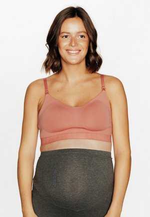 Femme enceinte aux cheveux bruns mi-longs portant un soutien-gorge de maternité rose et un pantalon gris foncé, souriante et face à la caméra.