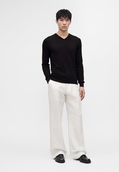 Jeune homme debout portant un pull noir à manches longues en V, un pantalon blanc ample et des chaussures noires sur un fond blanc uni.