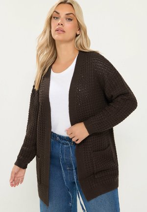 Vrouw met lang blond haar die een donkerbruine gebreide cardigan draagt over een wit overhemd en blauwe high-waist jeans tegen een effen achtergrond.