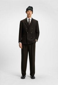 Abito doppio petto marrone scuro con revers, abbinato a una camicia bianca e un berretto nero a trama. Le scarpe nere completano l'outfit.