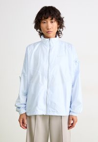 Persona con capelli ricci corti che indossa una giacca leggera azzurra con chiusura lampo e dettagli floreali decorativi sulle maniche, insieme a pantaloni beige a gamba larga.