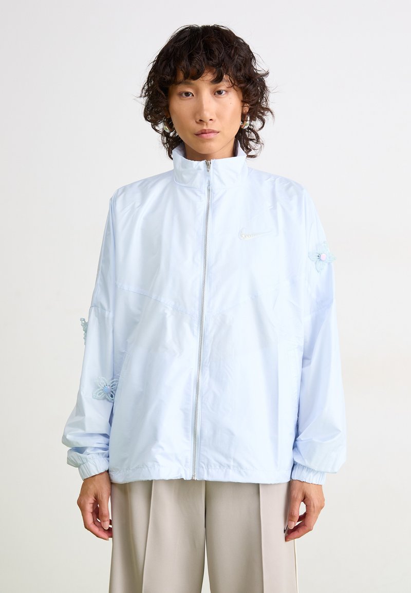 Persona con capelli ricci corti che indossa una giacca leggera azzurra con chiusura lampo e dettagli floreali decorativi sulle maniche, insieme a pantaloni beige a gamba larga.