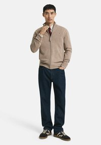 Man in een beige ritstrui, geruit shirt, rode stropdas, donkerblauwe jeans en zwarte sneakers met witte patronen, staand met één hand in zijn zak.