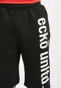 Shorts neri in felpa con una texture morbida, caratterizzati da un bold testo bianco "eckō unld." che corre in verticale lungo la gamba sinistra. Dettaglio con vita elastica.