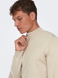 Beige Pullover mit hohem Kragen und einem halben Reißverschluss, gerippten Bündchen und glatter Textur. Das Model fasst den Kragen an und zeigt das eng anliegende Design.