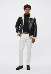 Czarna skórzana kurtka z podszewką z kożuszka, biały golf, dopasowane białe spodnie oraz czarne buty. Modelka stoi pewnie.