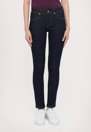 Osoba ubrana w ciemnoniebieskie obcisłe dżinsy, białe trampki i fioletową bluzkę, stojąca na tle jednolitego jasnego tła.