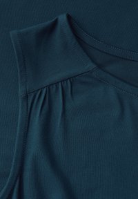 Tyrkysový bezrukávový top vyrobený z měkké látky; má na rameni zpevněný detail a hladký, strukturovaný povrch.