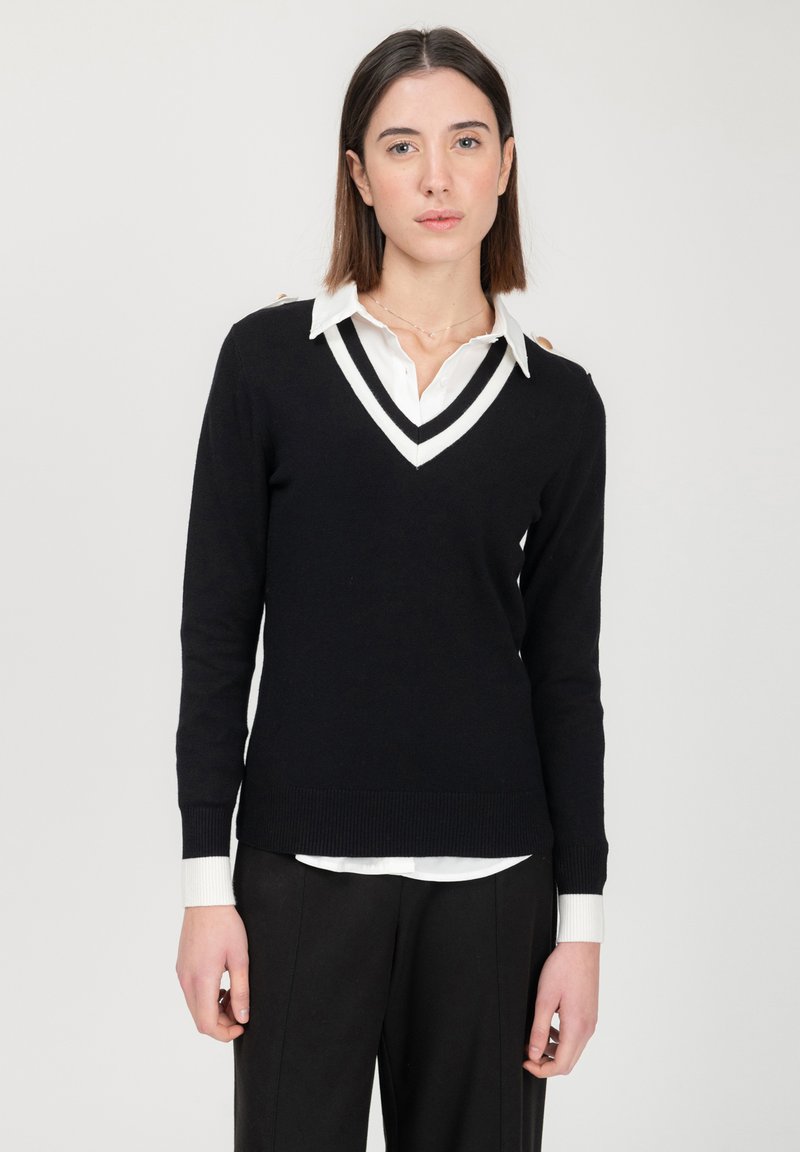 Schwarzer V-Ausschnitt-Pullover mit weißen Akzenten, mit einem gerippten Saum und Bündchen. Getragen über einem weißen Hemd mit Kragen, was einen geschichteten Look kreiert.