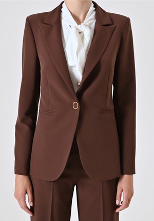 Blazer marron cintré avec un seul bouton doré, porté sur un chemisier blanc à col noué, assorti à un pantalon marron coordonné.