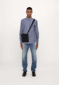 Borsa a tracolla nera con finitura testurizzata, indossata su una camicia a righe blu e bianche. Un paio di jeans blu chiaro a gamba dritta e scarpe nere completano il look.