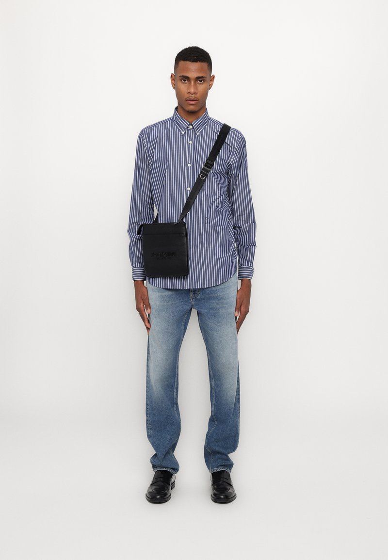 Borsa a tracolla nera con finitura testurizzata, indossata su una camicia a righe blu e bianche. Un paio di jeans blu chiaro a gamba dritta e scarpe nere completano il look.