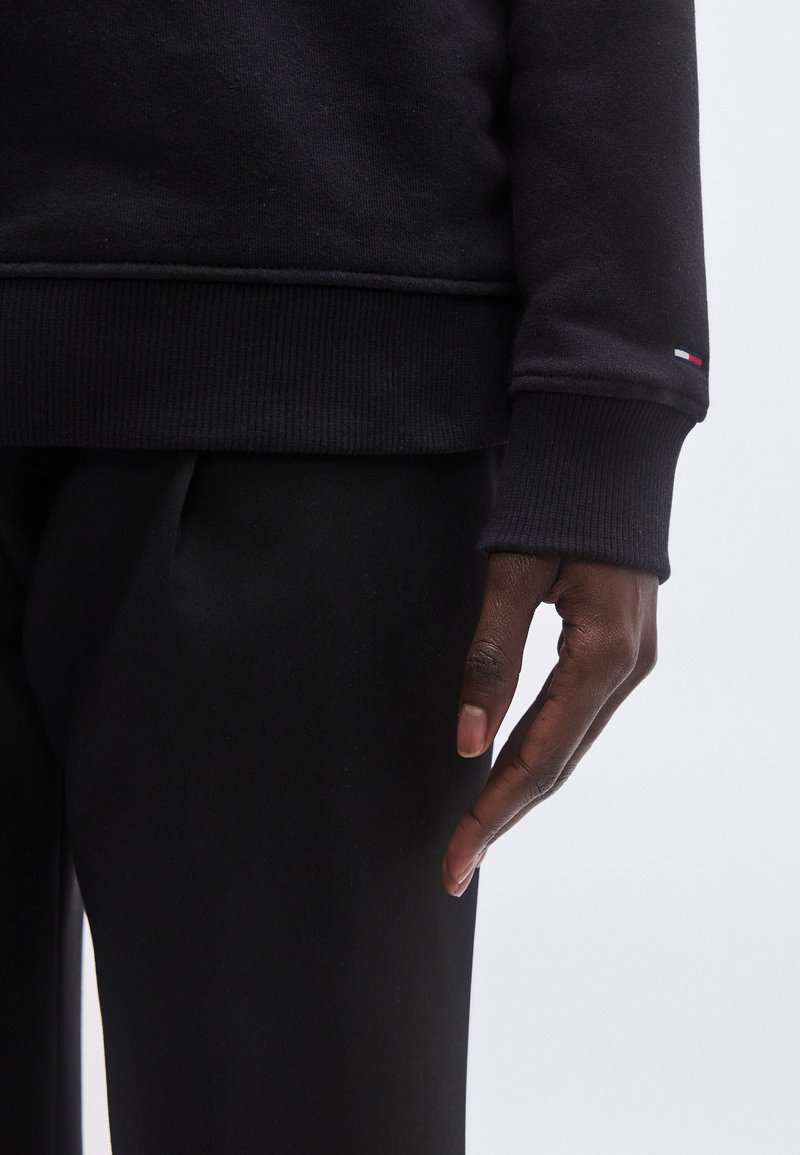 Black sweatshirt with ribbed cuffs, featuring a small tricolor logo on the sleeve. Worn with black pants, showing hand in a relaxed position.
