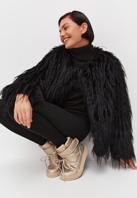 Giacca in finta pelliccia nera con una lunga trama shaggy, indossata sopra un dolcevita nero e leggings neri; abbinata a sneakers beige con lacci spessi.