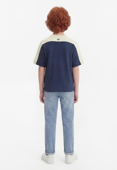 Kind met krullend rood haar staat met de rug naar de kijker, draagt een marineblauw-wit T-shirt met korte mouwen, lichtblauwe spijkerbroek en witte sneakers.