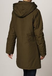 Personne portant un manteau long matelassé vert olive avec capuche, vue de dos, debout devant un fond clair uni.