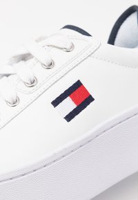 Baskets blanches avec bout rond, à lacets, semelle en caoutchouc et logo brodé rouge, blanc et bleu. Tige en cuir lisse avec des détails de couture.
