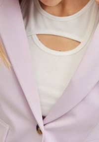 Liliowy żakiet noszony na białej bluzce z okrągłym dekoltem z wycięciami. Żakiet ma zapięcie na pojedynczy guzik i gładką fakturę.