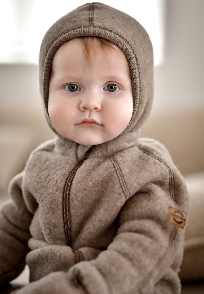 Brauner Fleece-Schneeanzug für Säuglinge mit Reißverschluss, angenähter Kapuze und kontrastierenden Nähten. Verfügt über ein Logo-Patch am Ärmel. Weiche Textur.