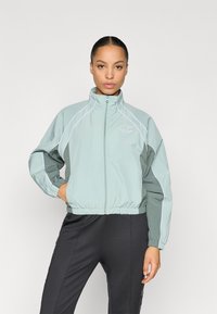 Ελαφρύ, μίντ πράσινο αδιάβροχο μπουφάν με ψηλό γιακά, ελαστική μέση και αντανακλαστικές λευκές λεπτομέρειες. Το λογότυπο εμφανίζεται στο στήθος.