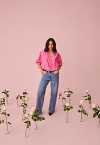 Femme en blouse rose et jean se tient parmi des roses roses dans des vases en verre sur un sol et un fond roses.