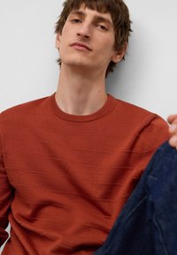 Giovane uomo con capelli corti castani che indossa un maglione testurizzato color ruggine e jeans blu scuro, seduto su uno sfondo bianco.
