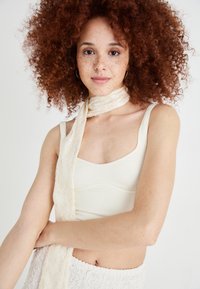Cream-colored ribbed bralette with adjustable straps, paired with a cream lace scarf. Model has curly red hair and light freckles.