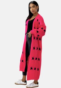 Lang åbent strikket cardigan i lys pink med sorte stjernemønstre. Designet inkluderer ribbet kant og lange ærmer.