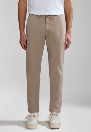 Mężczyzna w beżowych spodniach chino i białych trampkach stoi na jednolitym jasnoszarym tle.