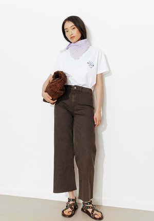 Femme portant un t-shirt blanc graphique avec un foulard lavande autour du cou, pantalon large noir, sandales cloutées, tenant une pochette marron imprimé léopard.