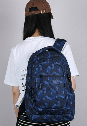 Personne aux longs cheveux noirs portant une casquette marron et une chemise blanche, portant un grand sac à dos bleu et noir à motifs, vue de dos.