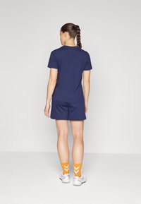 Navyblaue Baumwoll-T-Shirt und Shorts, mit orangenen Socken, die ein weißes Chevron-Muster aufweisen. Das Modell wird von hinten betrachtet.