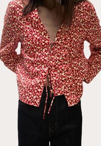 Rød blomstermønstret bluse med dyb V-hals, knyttet frontdetalje og lange ærmer, lavet af letvægtsstof. Sammen med mørke jeans.