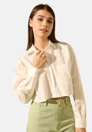 Jeune femme aux longs cheveux bruns portant une chemise blanche boutonnée loose et courte ainsi qu'un pantalon vert clair taille haute, posant avec confiance.