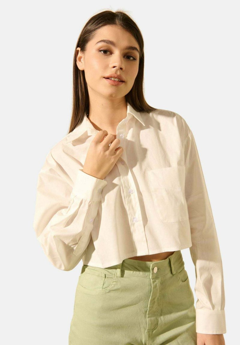 Jeune femme aux longs cheveux bruns portant une chemise blanche boutonnée loose et courte ainsi qu'un pantalon vert clair taille haute, posant avec confiance.